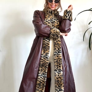 May include: A long, brown leather coat with leopard print trim and accents. The coat features a high collar and is worn over a leopard print top. The coat is floor-length and has a classic, vintage style.