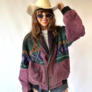 Könnte beinhalten: Eine mehrfarbige Wildleder-Bomberjacke mit dunkelvioletter Basis und grün-schwarzen Mustereinsätzen an Brust und Rücken. Die Jacke verfügt über einen schwarzen Rippstrickkragen, Rippbündchen an Ärmeln und Saum sowie eine Knopfleiste und Seitentaschen.