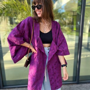 Puede incluir: Una chaqueta abierta de color morado vibrante con un patrón de hojas tonal texturizado, mangas anchas de tres cuartos y un corte relajado. La prenda se lleva sobre un top corto negro y vaqueros azul claro, junto con un pequeño bolso bandolera marrón.