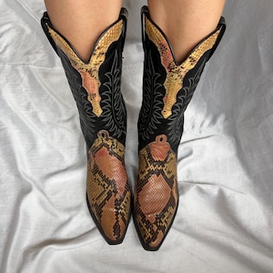 Puede incluir: Un par de botas de vaquero negras con un estampado de piel de serpiente en el pie y el cuello superior. Las botas tienen una punta puntiaguda y costuras decorativas. El estampado de piel de serpiente presenta tonos marrones, beige y amarillos.