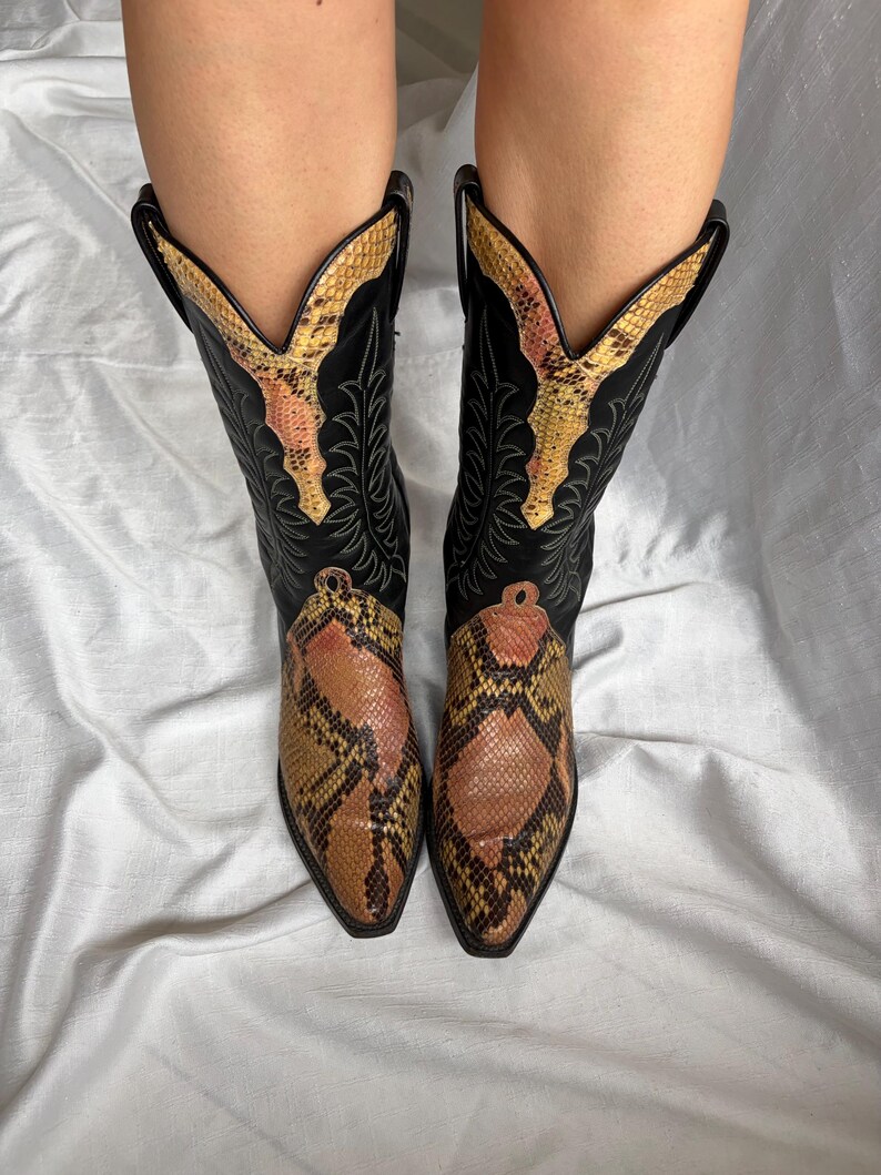Puede incluir: Un par de botas de vaquero negras con un estampado de piel de serpiente en el pie y el cuello superior. Las botas tienen una punta puntiaguda y costuras decorativas. El estampado de piel de serpiente presenta tonos marrones, beige y amarillos.