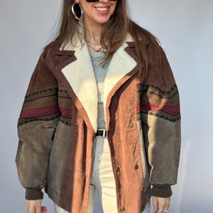 Peut inclure: Veste en daim marron avec col et doublure en fausse fourrure crème. La veste présente un motif géométrique rouge, vert et noir sur les manches. Elle a une coupe croisée avec des boutons dorés et une ceinture.