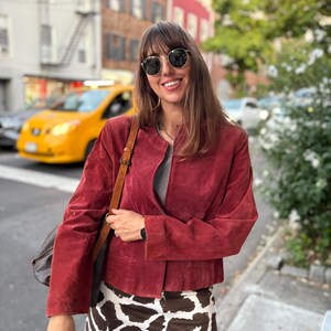May include: A woman wearing a red suede jacket, a gray top, and a cow print mini skirt. She is also wearing sunglasses and carrying a brown leather bag. The background shows a city street with buildings and a yellow taxi.