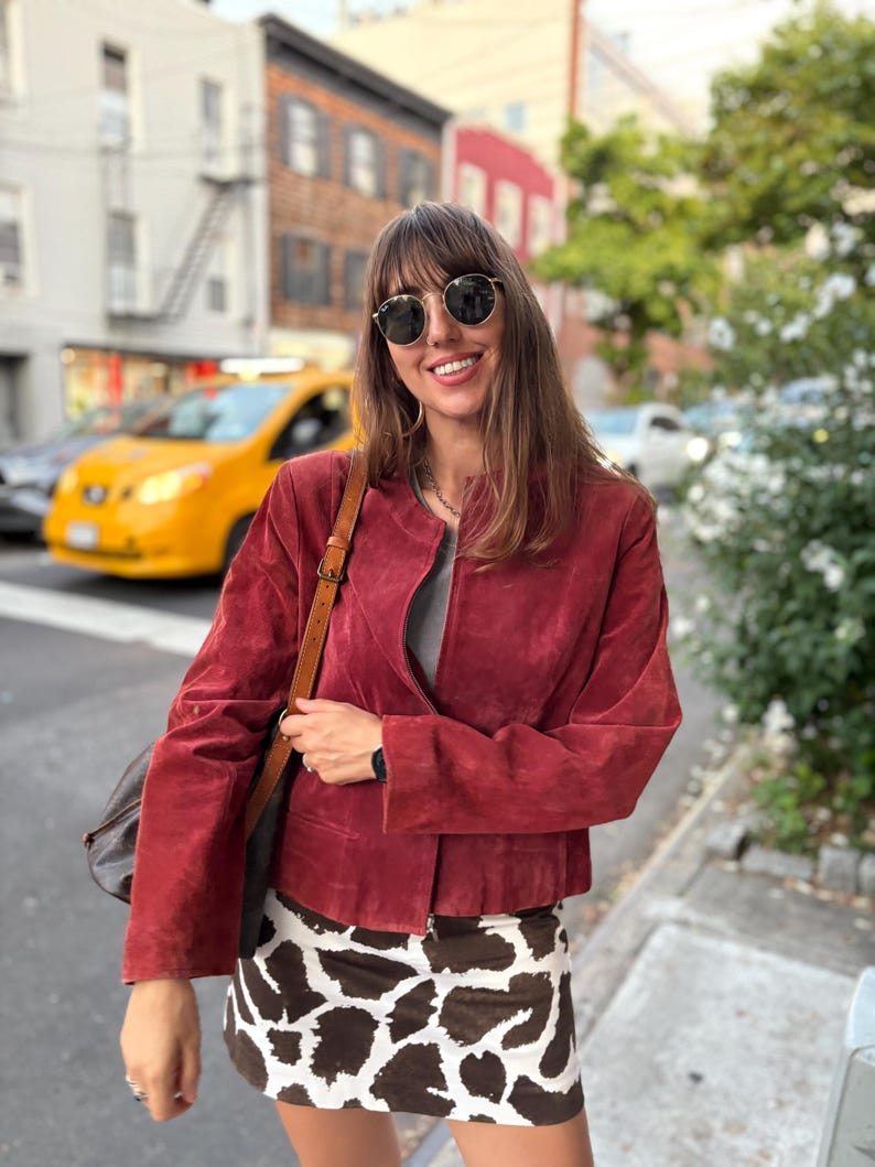 May include: A woman wearing a red suede jacket, a gray top, and a cow print mini skirt. She is also wearing sunglasses and carrying a brown leather bag. The background shows a city street with buildings and a yellow taxi.
