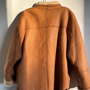 May include: A brown leather jacket with a shearling collar and lining, hanging on a metal hanger. The jacket has a classic design with button closures and detailed stitching. The jacket's exterior is a warm, medium brown color.