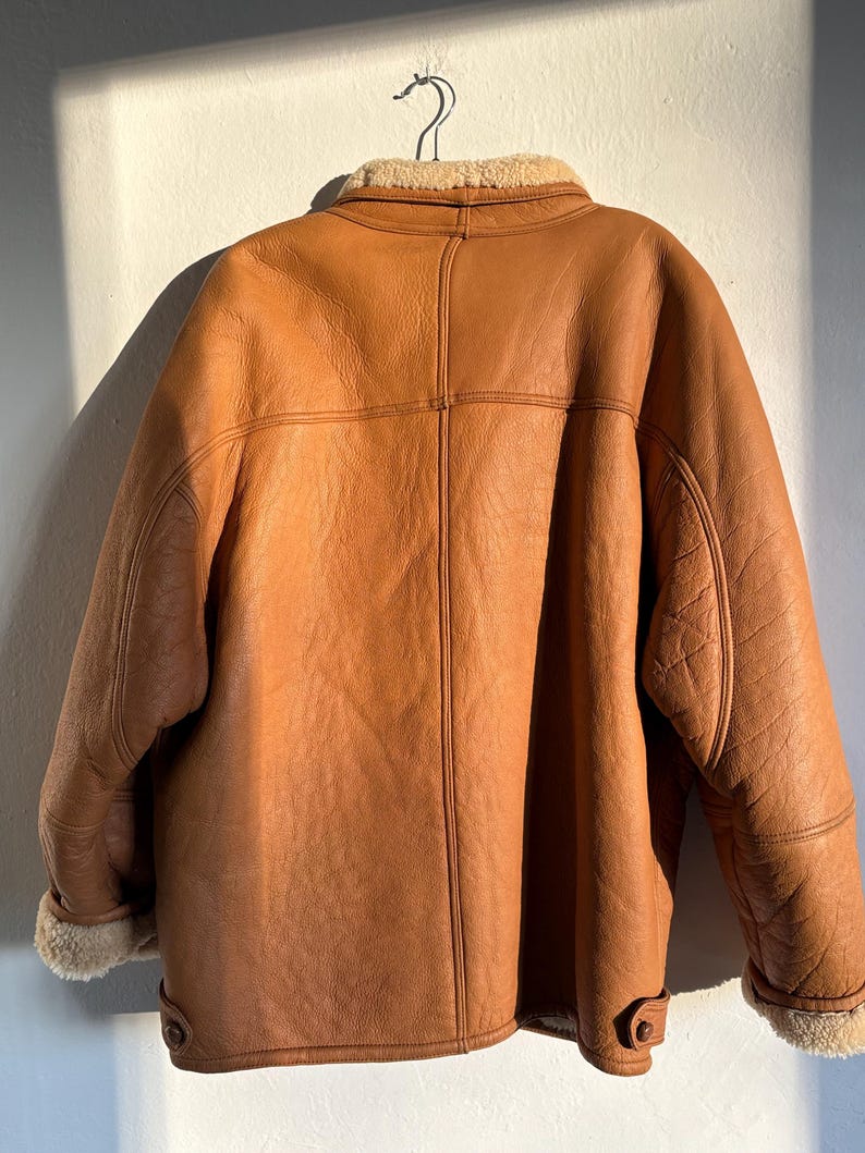 May include: A brown leather jacket with a shearling collar and lining, hanging on a metal hanger. The jacket has a classic design with button closures and detailed stitching. The jacket's exterior is a warm, medium brown color.