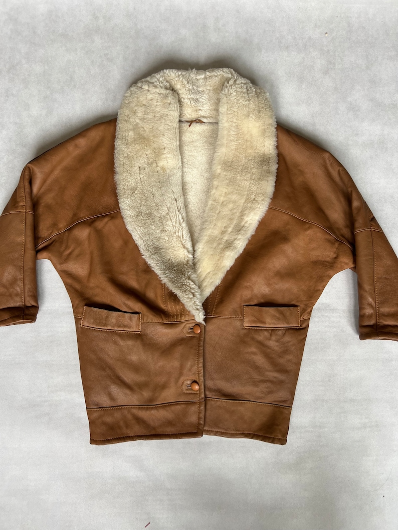 May include: A brown leather jacket with a large white faux fur collar. The jacket has a button closure and two pockets.