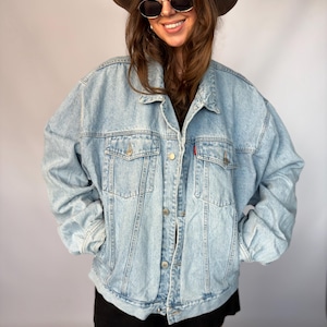 May include: An oversized light blue denim jacket featuring a classic collar, long sleeves, metal button closures, and two chest pockets with buttoned flaps. The jacket has a relaxed fit with visible stitching details and a small red brand tab.