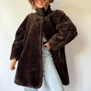 Può includere: Un cappotto in pelliccia sintetica marrone scuro con collo alto e chiusura a alamaro. Il cappotto presenta maniche lunghe e un orlo dritto. È indossato con jeans azzurri, un cappello a tesa larga marrone e grandi occhiali da sole colorati.