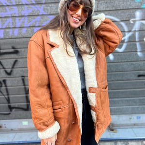 Puede incluir: Una chaqueta de cuero color canela con un grueso forro de borrego color crema, con solapa ancha, mangas largas con puños de borrego y tres botones frontales. Se lleva sobre una camiseta gris y pantalones negros, junto con un gorro de punto beige.