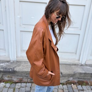 May include: A tan leather blazer featuring a notched lapel, long sleeves, and two front buttons. The jacket has a structured shoulder design and two side pockets. It is worn over a white top and paired with light blue denim jeans.