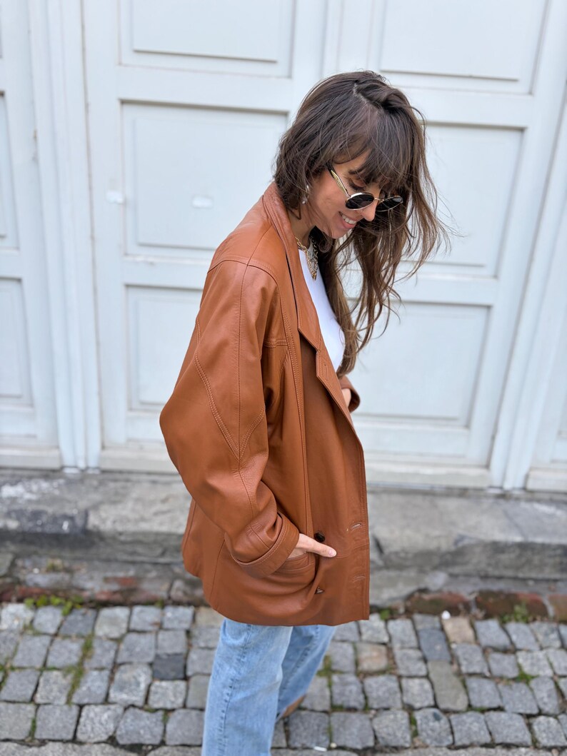 May include: A tan leather blazer featuring a notched lapel, long sleeves, and two front buttons. The jacket has a structured shoulder design and two side pockets. It is worn over a white top and paired with light blue denim jeans.