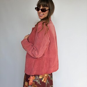 May include: A dusty rose suede blazer with long sleeves, a collared neckline, and front pockets, paired with a dark burgundy skirt featuring a vibrant floral print in shades of orange, yellow, and green. The blazer has a buttoned front closure.