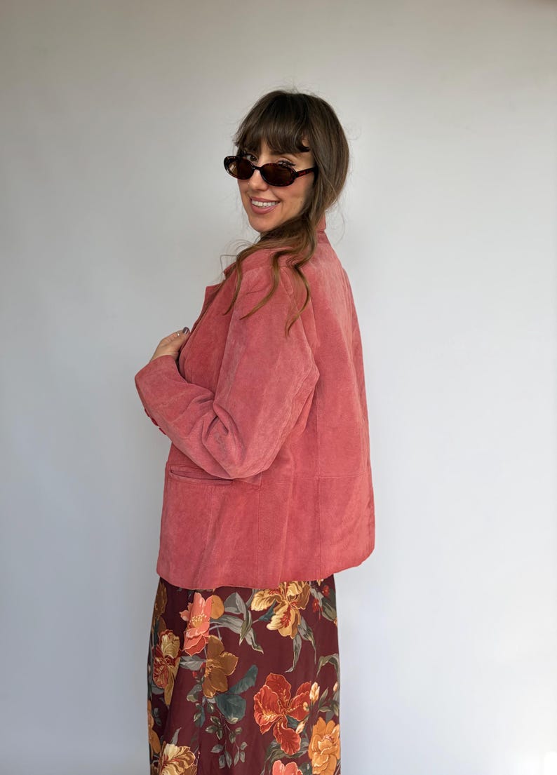May include: A dusty rose suede blazer with long sleeves, a collared neckline, and front pockets, paired with a dark burgundy skirt featuring a vibrant floral print in shades of orange, yellow, and green. The blazer has a buttoned front closure.
