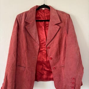 May include: A coral-colored blazer with a classic notched lapel and three-button closure. The blazer features a matching red satin lining and three buttons on each cuff. The jacket is hanging on a black hanger.