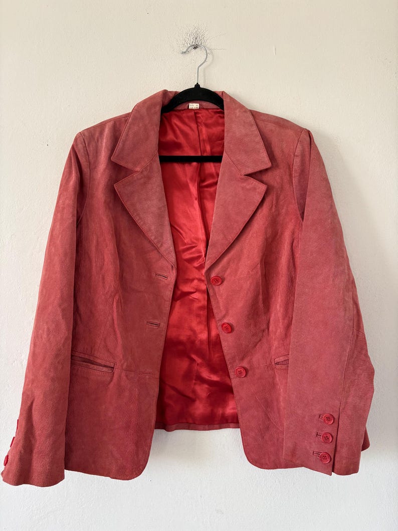 May include: A coral-colored blazer with a classic notched lapel and three-button closure. The blazer features a matching red satin lining and three buttons on each cuff. The jacket is hanging on a black hanger.