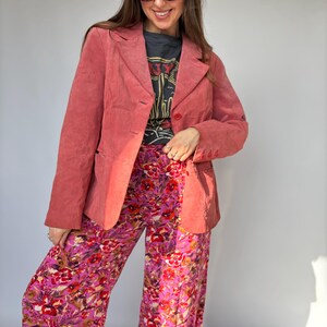 May include: A coral-colored suede blazer with a notched lapel and button closure, worn over a dark gray graphic t-shirt and wide-leg trousers featuring a vibrant pink, purple, and red floral pattern with green leaf accents.