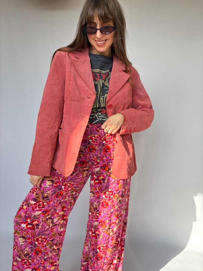 May include: A coral-colored suede blazer with a notched lapel and button closure, worn over a dark gray graphic t-shirt and wide-leg trousers featuring a vibrant pink, purple, and red floral pattern with green leaf accents.