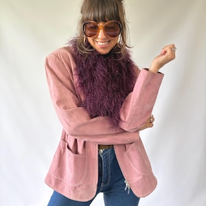 May include: A dusty pink suede jacket featuring a deep purple textured faux fur collar, long sleeves, and two front patch pockets. The jacket is worn with blue denim jeans and large brown-tinted oversized sunglasses with thick frames.