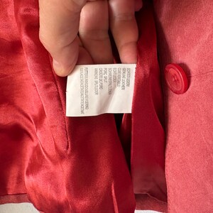 May include: A red suede jacket with a matching red button and a shiny red acetate lining. A white care label is sewn into the interior, featuring text in multiple languages including Genuine Leather, Porc Split, and 100% Acetate.