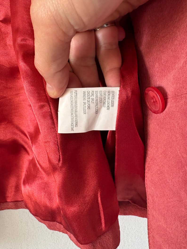 May include: A red suede jacket with a matching red button and a shiny red acetate lining. A white care label is sewn into the interior, featuring text in multiple languages including Genuine Leather, Porc Split, and 100% Acetate.
