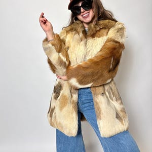 Puede incluir: Un abrigo de piel vintage en tonos marrón, beige y crema. El abrigo tiene una silueta larga y entallada con cierre de botón. La persona lleva una gorra de béisbol marrón y vaqueros azules.