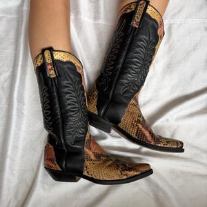 Puede incluir: Botas de vaquero con cuero negro y detalles de estampado de serpiente. Las botas tienen costuras decorativas y una puntera puntiaguda. El estampado de serpiente presenta tonos marrones, beige y naranja. Sobre un fondo blanco.