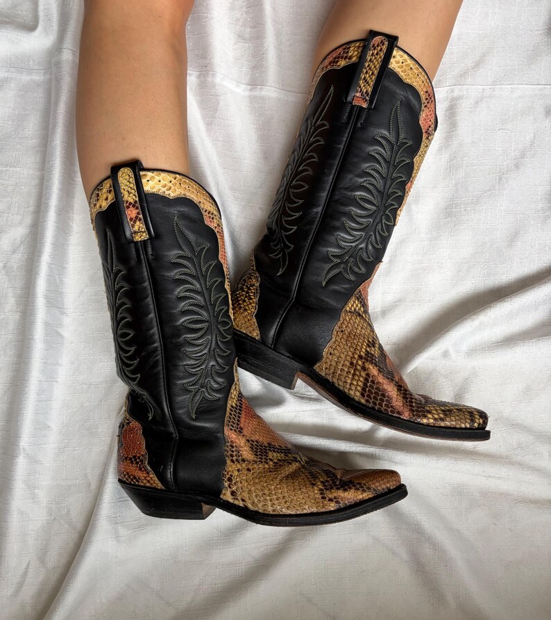 Puede incluir: Botas de vaquero con cuero negro y detalles de estampado de serpiente. Las botas tienen costuras decorativas y una puntera puntiaguda. El estampado de serpiente presenta tonos marrones, beige y naranja. Sobre un fondo blanco.