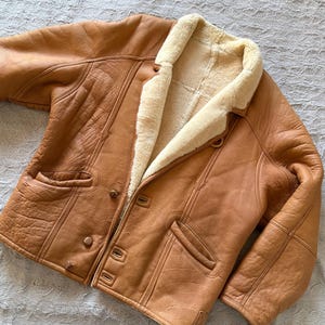 May include: A tan leather jacket with a cream-colored shearling lining. The jacket has a button-up front, two front pockets, and a notched collar. The leather has a slightly distressed appearance. The jacket is a classic outerwear piece.