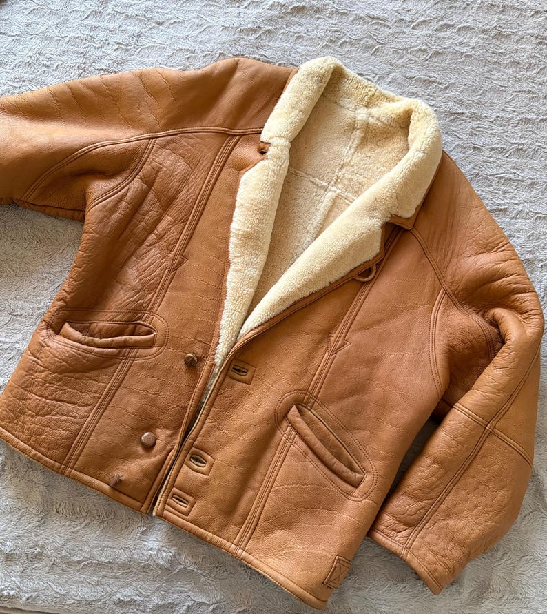 May include: A tan leather jacket with a cream-colored shearling lining. The jacket has a button-up front, two front pockets, and a notched collar. The leather has a slightly distressed appearance. The jacket is a classic outerwear piece.