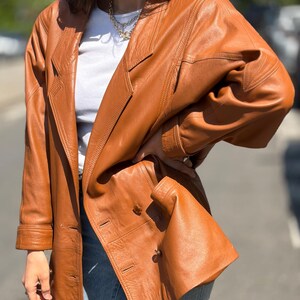 May include: A tan leather blazer featuring a double-breasted front with visible buttons, notched lapels, and long sleeves with cuffs. The jacket has structured shoulders and a smooth finish, worn over a white top and light blue denim jeans.