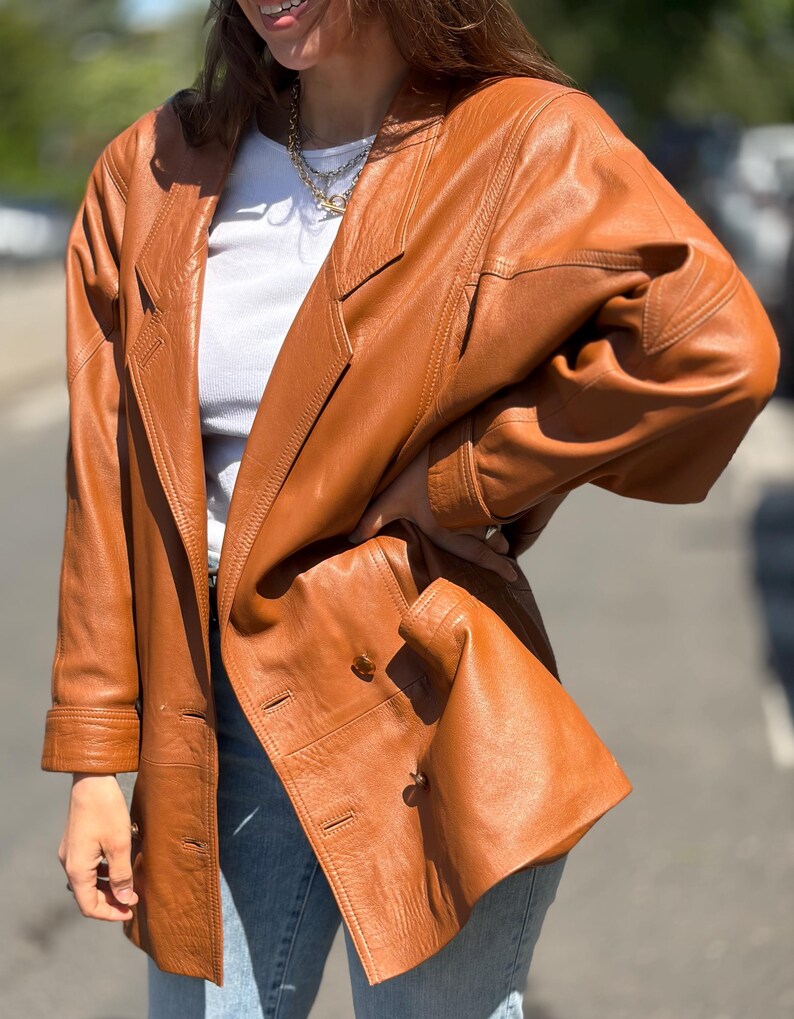 May include: A tan leather blazer featuring a double-breasted front with visible buttons, notched lapels, and long sleeves with cuffs. The jacket has structured shoulders and a smooth finish, worn over a white top and light blue denim jeans.