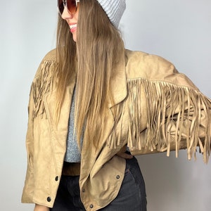 Könnte beinhalten: Eine braune Lederjacke mit Fransen. Die Jacke hat einen Reißverschluss und einen Stehkragen. Das Model trägt eine weiße Mütze und eine Sonnenbrille.