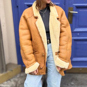 May include: A tan leather jacket with a cream-colored shearling lining and trim. The jacket has a classic design with a notched collar, button closures, and side pockets. The jacket is worn over a dark gray shirt and light blue jeans.