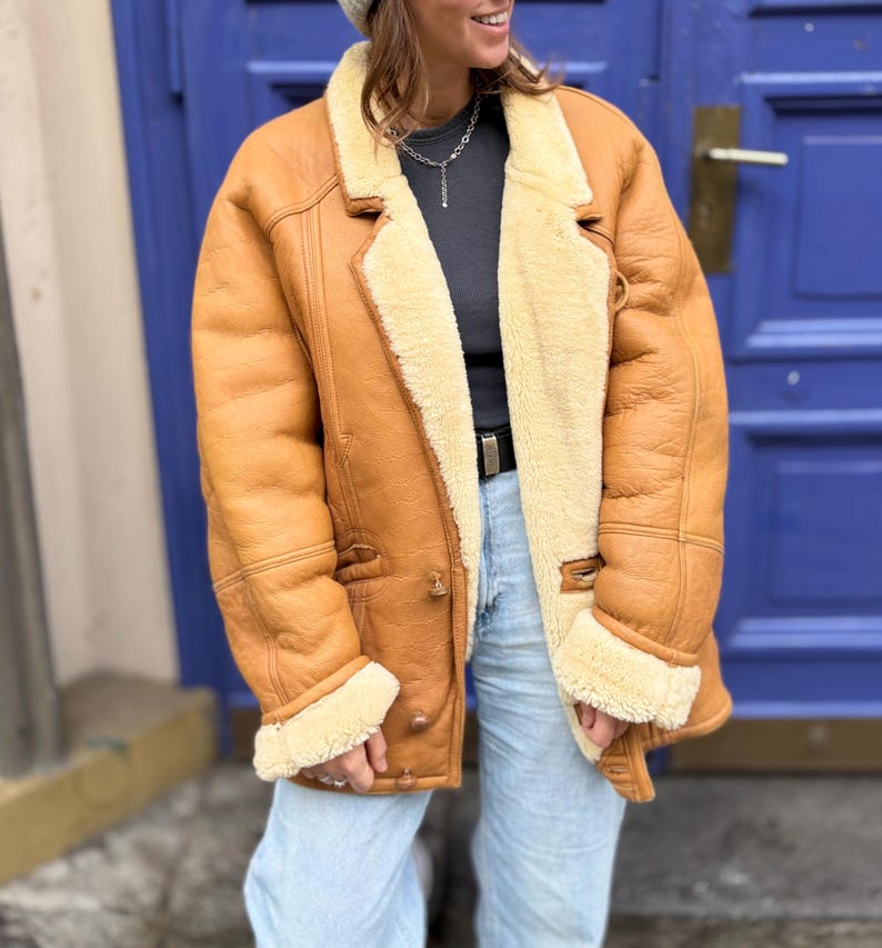 May include: A tan leather jacket with a cream-colored shearling lining and trim. The jacket has a classic design with a notched collar, button closures, and side pockets. The jacket is worn over a dark gray shirt and light blue jeans.