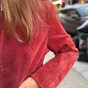 May include: A red suede jacket with a silver zipper. The jacket has long sleeves and a classic design. The image shows the jacket's texture and color, with a hand in the pocket.