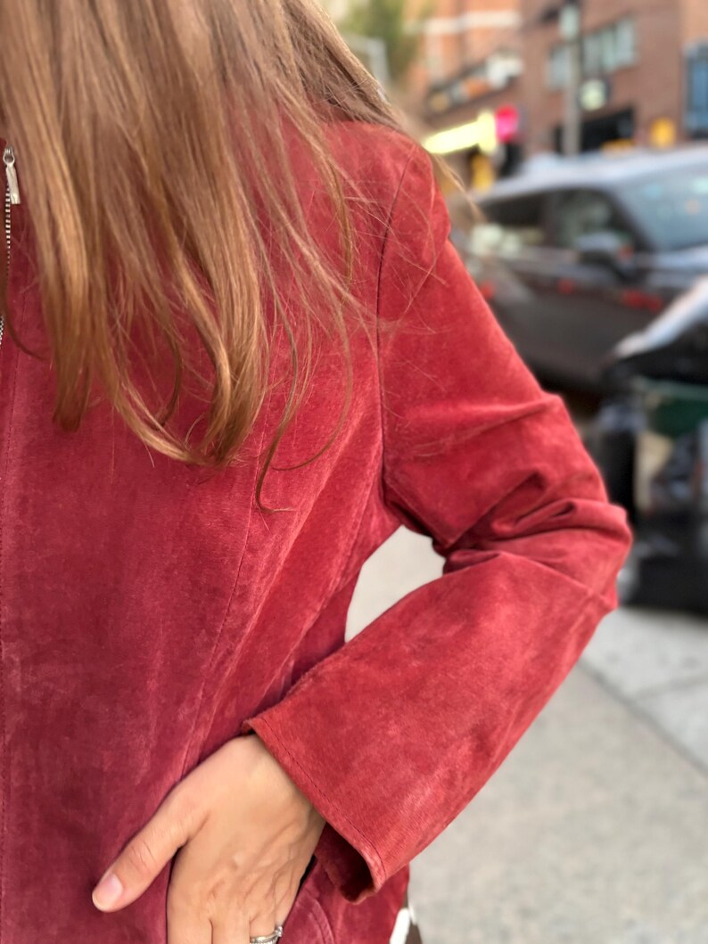 May include: A red suede jacket with a silver zipper. The jacket has long sleeves and a classic design. The image shows the jacket's texture and color, with a hand in the pocket.