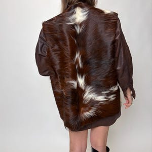 May include: A brown and white cowhide jacket with brown leather sleeves. The jacket is worn with a brown beret and black boots. The back of the jacket is the focal point, showcasing the natural fur pattern.