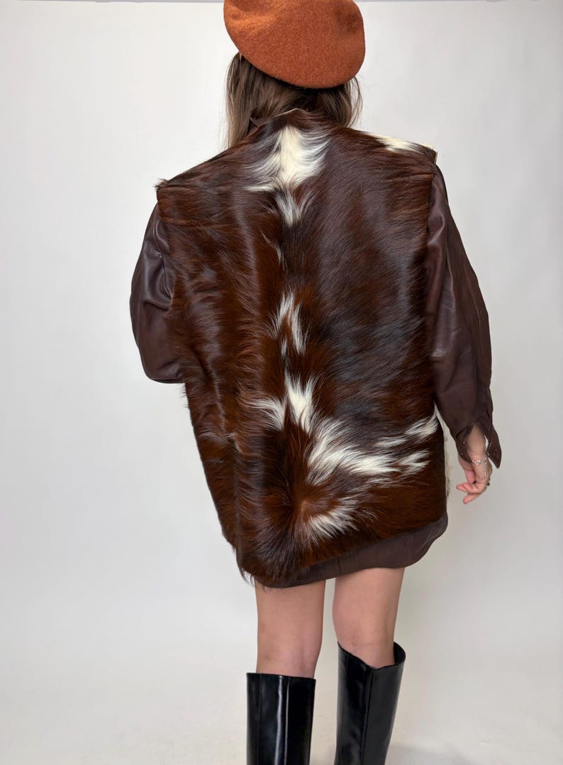 May include: A brown and white cowhide jacket with brown leather sleeves. The jacket is worn with a brown beret and black boots. The back of the jacket is the focal point, showcasing the natural fur pattern.