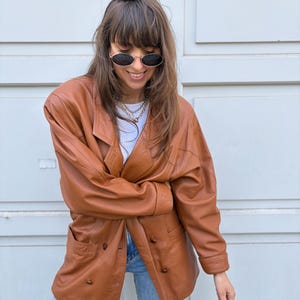 May include: A tan leather blazer featuring a double-breasted front with visible buttons, long sleeves, and a wide lapel collar. The jacket is paired with a white top and light blue denim jeans, complemented by oval-shaped sunglasses and a layered chain necklace.