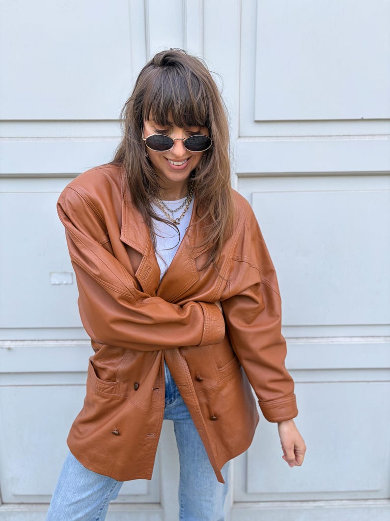 May include: A tan leather blazer featuring a double-breasted front with visible buttons, long sleeves, and a wide lapel collar. The jacket is paired with a white top and light blue denim jeans, complemented by oval-shaped sunglasses and a layered chain necklace.