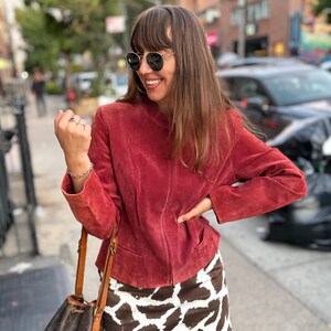 May include: A red suede jacket with a zip-up front and a brown and white cow print mini skirt. The woman is wearing sunglasses and carrying a brown leather handbag.