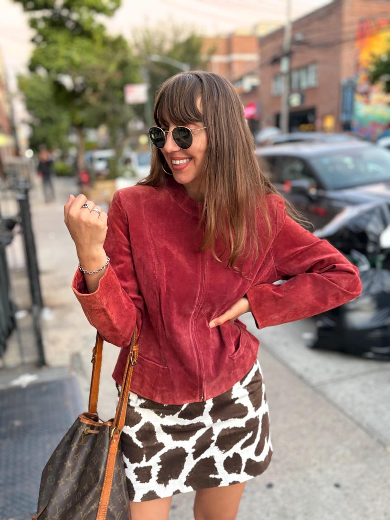May include: A red suede jacket with a zip-up front and a brown and white cow print mini skirt. The woman is wearing sunglasses and carrying a brown leather handbag.