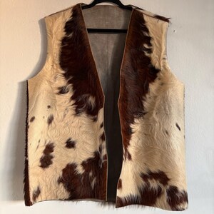 May include: A sleeveless vest made of cowhide. The vest features a cream-colored base with irregular patches of dark brown fur. The vest is hanging on a black hanger against a white wall.