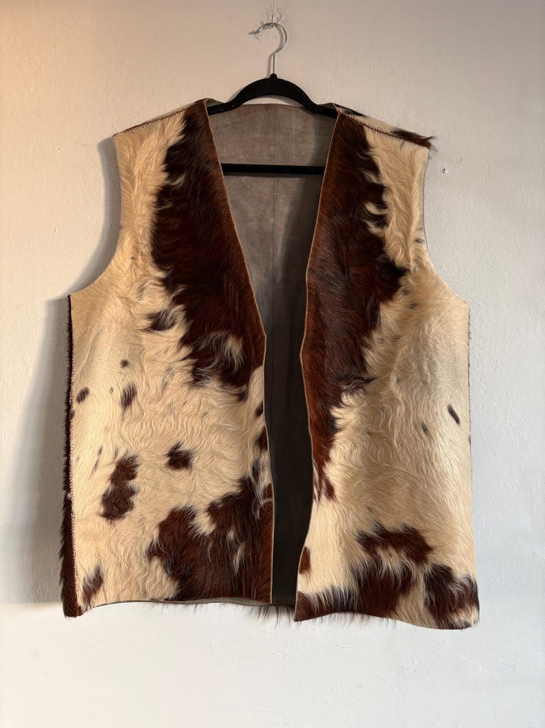 May include: A sleeveless vest made of cowhide. The vest features a cream-colored base with irregular patches of dark brown fur. The vest is hanging on a black hanger against a white wall.