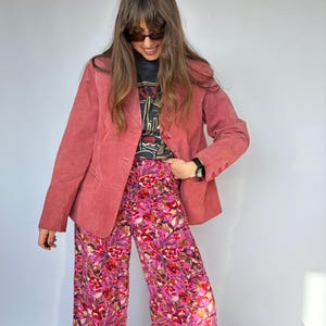 May include: A dusty rose blazer with a three-button closure is paired with wide-leg floral pants in shades of pink and purple. The outfit includes a black graphic tee. The ensemble is a stylish, vintage-inspired look.