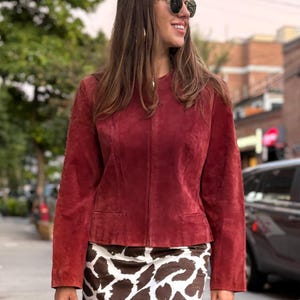 May include: A burgundy suede jacket with a round collar and two front pockets. The jacket is paired with a brown and white patterned mini skirt. The outfit is worn outdoors on a sunny day.