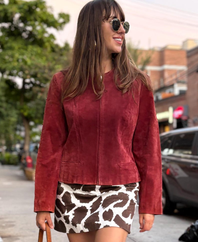 May include: A burgundy suede jacket with a round collar and two front pockets. The jacket is paired with a brown and white patterned mini skirt. The outfit is worn outdoors on a sunny day.