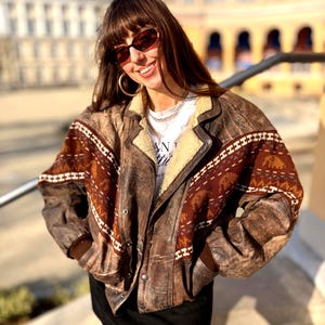 Puede incluir: Una chaqueta bomber de cuero marrón con cuello de piel sintética color crema y una banda horizontal ancha en el pecho y las mangas con un patrón geométrico en rojo oscuro y blanco. La chaqueta tiene mangas largas con puños de canalé marrones.