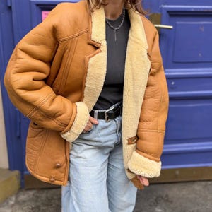 May include: A brown shearling jacket with a cream-colored shearling lining and trim. The jacket has a classic lapel collar, button closures, and a belted waist. The jacket is worn with a black belt and light blue jeans.